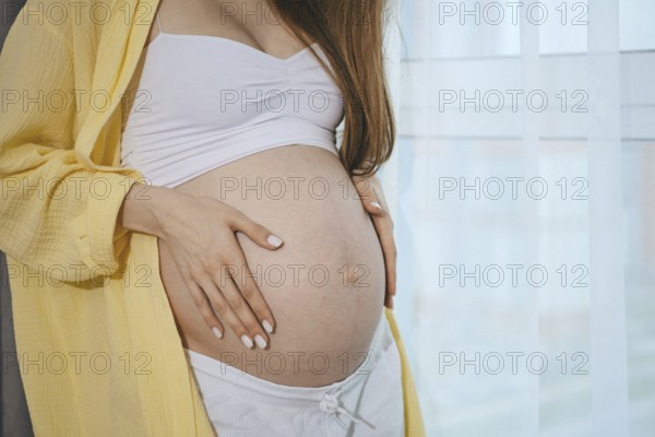 Close-up view of a baby bump of pregnant woman by the window. A woman lovingly places her hands on