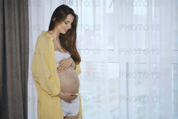 A pregnant woman stands by a window, smiling as she gently cradles her baby bump. Soft morning