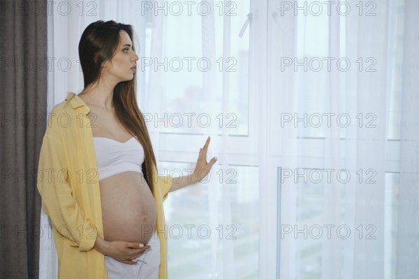 A pregnant woman stands by a window in a contemporary apartment, wearing a white top and loose