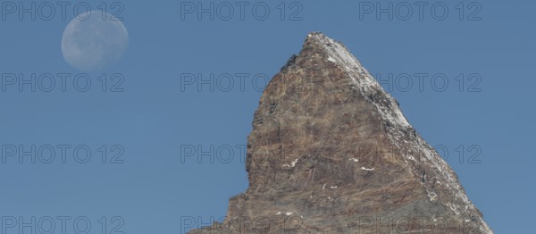 The Matterhorn mountain peak rises steeply against a bright blue sky. The full moon glistens gently