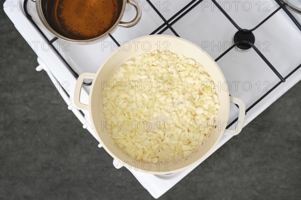 Chopped onions cooking in a white pan over medium heat on a stove. A pot of simmering broth sits