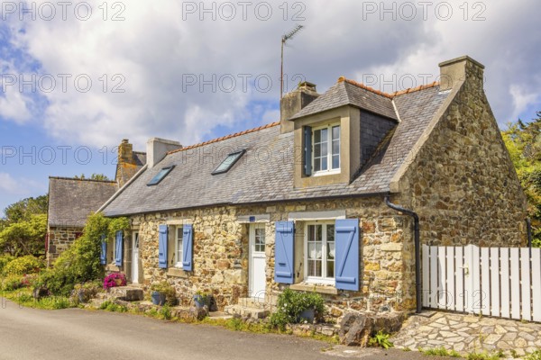 Old idyllic stone cottage with blue shutters and blooming flowers at the house wall by a road in