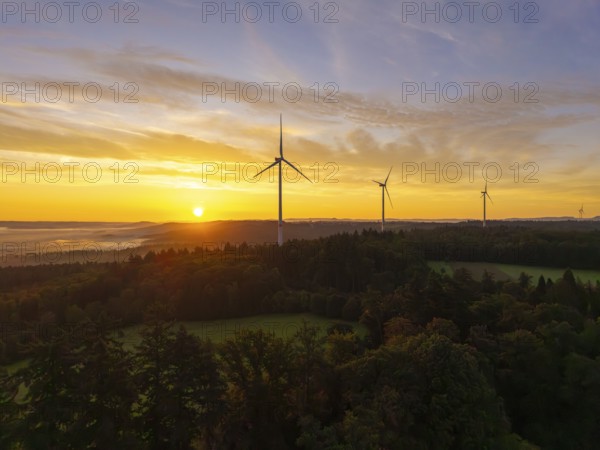 Wind turbines in a hilly and wooded landscape, illuminated by the rising sun, near Schorndorf,