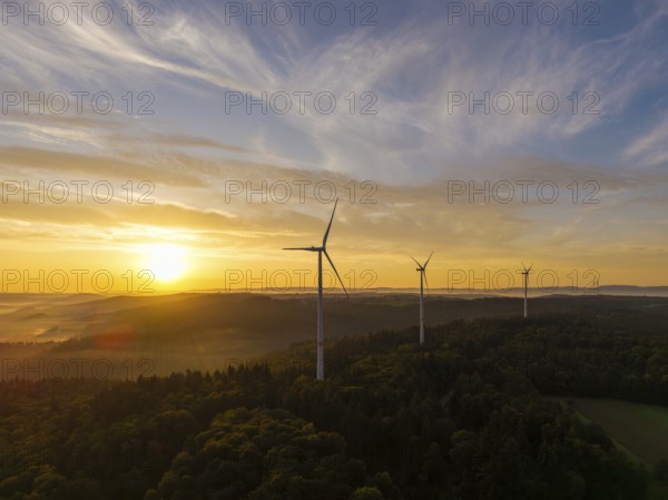Wind turbines at dusk over wooded hills, the light gently breaking through the clouds, near