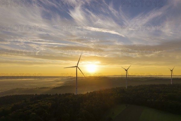 Wind turbines stand still under a soft sunlight illuminating the sky and forest, near Schorndorf,
