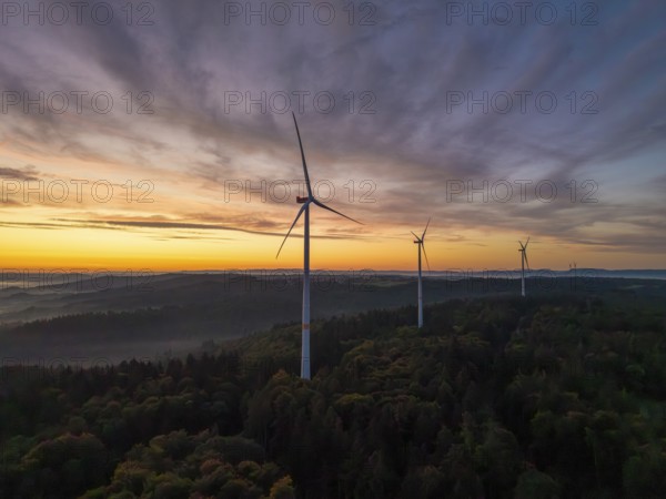 Wind turbines stand in a wooded landscape under a pastel-coloured sky at sunrise, near Schorndorf,