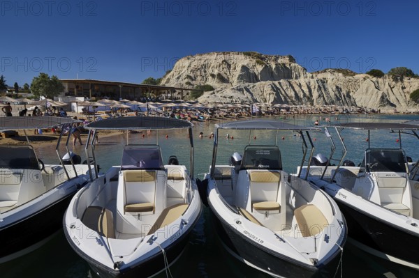Several boats are moored on a jetty with a rocky beach in the background. Holidaymakers relaxing on