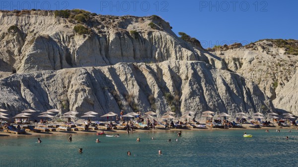 Beach in front of a large rock face, lined with sun loungers and umbrellas, bathers enjoying the