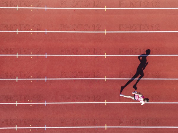 Aerial view of a female individual (runner) on a red athletics track (tartan track) with shadow as