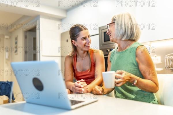 Two women sitting at a kitchen counter, engaging in conversation while using a laptop, one enjoying