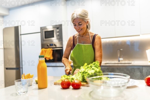 Happy mature woman wearing green apron cutting lettuce while preparing healthy salad in modern
