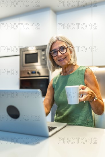 Smiling senior woman enjoying coffee while working on her laptop in a cozy kitchen, embracing a