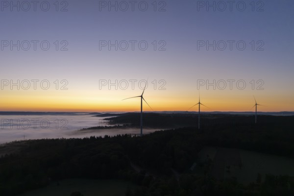 Wind turbines in front of a bright blue and orange sky at sunrise with fog over the landscape, near