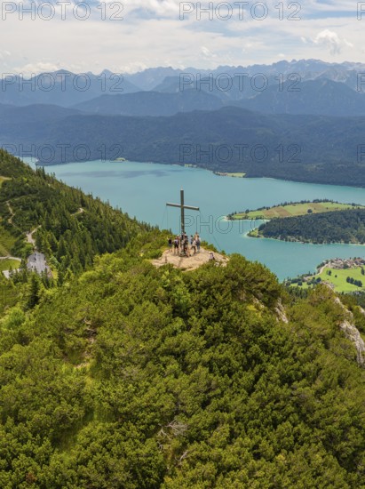 Group on a mountain with summit cross, overlooking a wide lake landscape, Herzogstand summit,