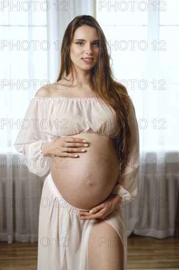 A pregnant woman stands gracefully in a softly lit room, wearing a flowing dress. She gently