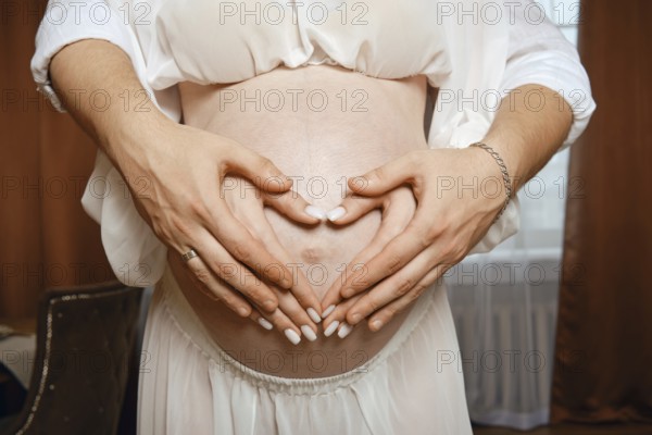 Two hands form a heart shape over a pregnant belly in a warm, softly lit room. The parents express