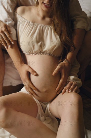 A couple sits on a soft bed, with one partner gently cradling the pregnant belly. The intimate