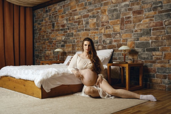 A pregnant woman sits elegantly in a warm-toned bedroom, showcasing her baby bump. The room
