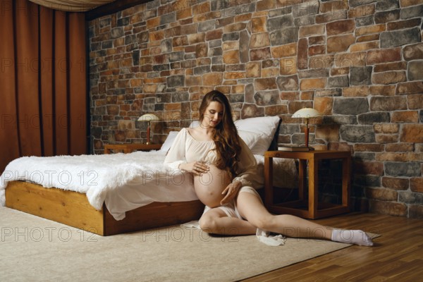 A pregnant woman sits in a warm, inviting bedroom, gently cradling her baby bump. The space