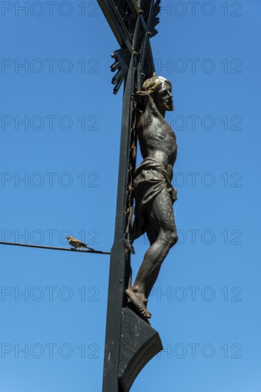 A detailed sculpture depicting Jesus on a cross rises against a bright blue sky. A small bird can