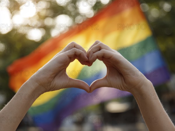 Two hands form a heart in front of a waving rainbow flag in the sunlight, symbolising equality,