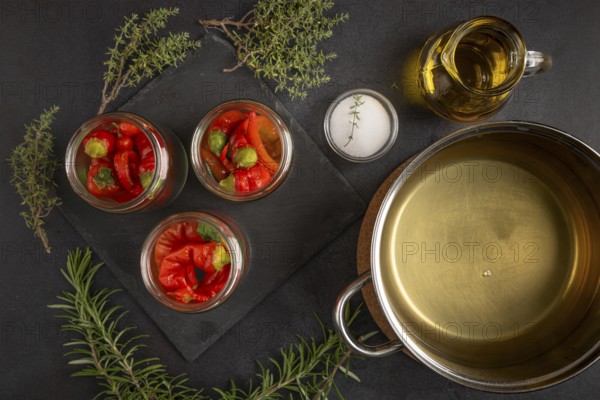 Glasses filled with chilli peppers next to a saucepan with vinegar, oil and herbs on a dark