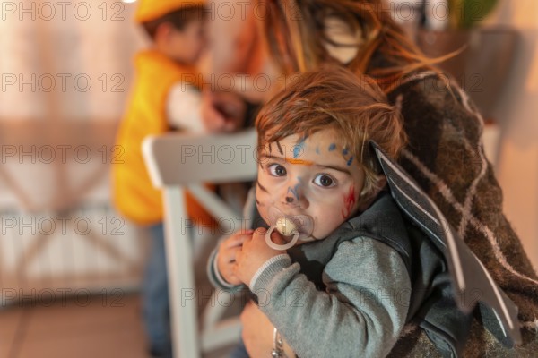 Toddler wearing a halloween costume with painted face and pacifier, held by mother, with another
