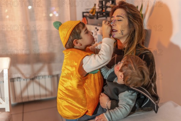 Two young brothers in halloween costumes playfully applying makeup on their mother's face in a