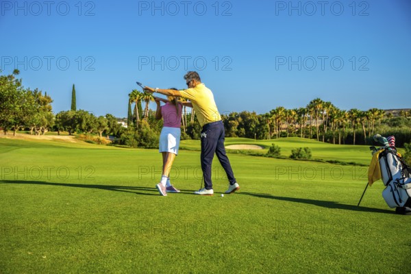 Middle aged golf instructor assisting young woman golfer on improving her swing on a sunny day at