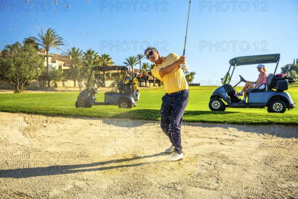 Male golfer hitting ball out of sand trap with golf cart and other players in background on sunny