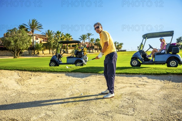 Golfer focuses on a bunker shot while others relax in golf carts on a bright, sunny day at a lush