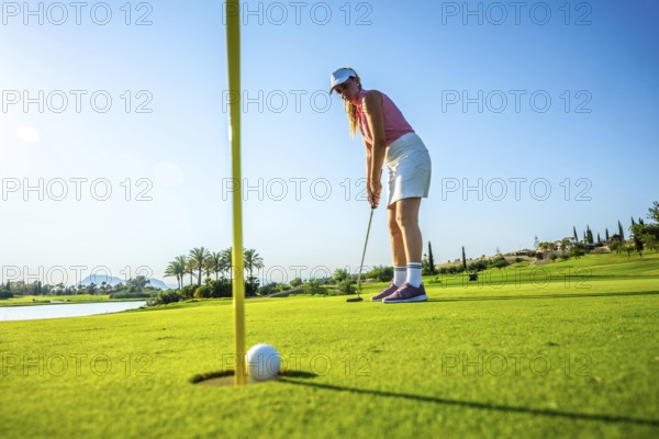 Woman focusing on a putt on a bright, sunny day at a lush golf course, with a ball near the hole