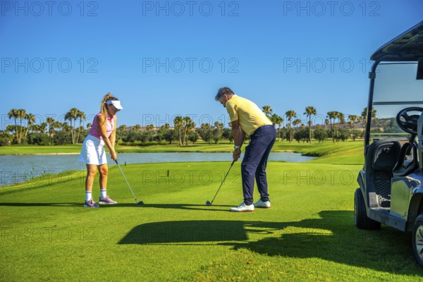 Father and daughter duo playing golf on a sunny day, surrounded by palm trees and a clear blue sky,
