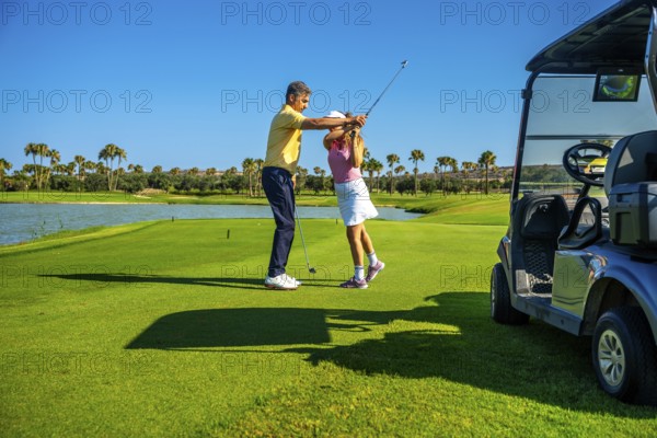 Golf instructor guiding female golfer on a sunny course, improving her swing technique near a golf
