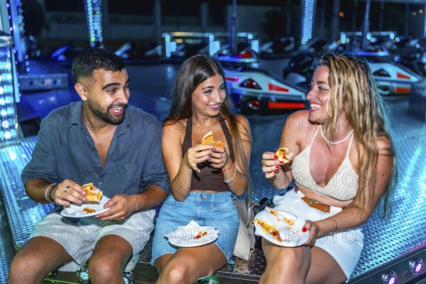 Three friends are enjoying sandwiches while sitting on a bumper cars platform, illuminated by
