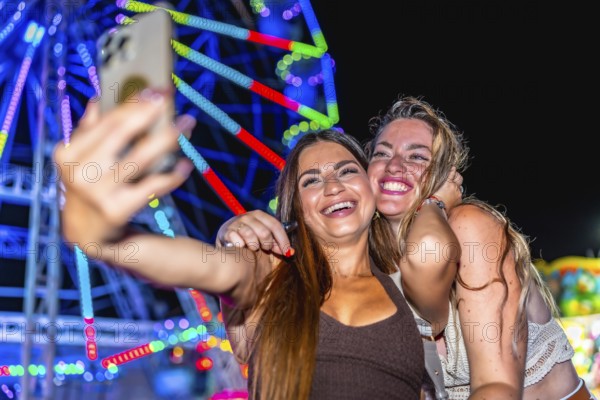 Two cheerful young women taking a selfie in front of a brightly lit ferris wheel at an amusement