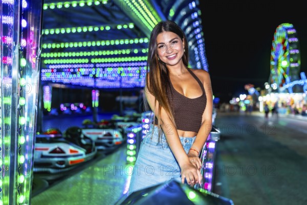Happy young woman posing joyfully near vibrant, illuminated bumper cars at an amusement park,