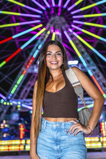 Happy young woman posing at night with illuminated ferris wheel in background, enjoying funfair