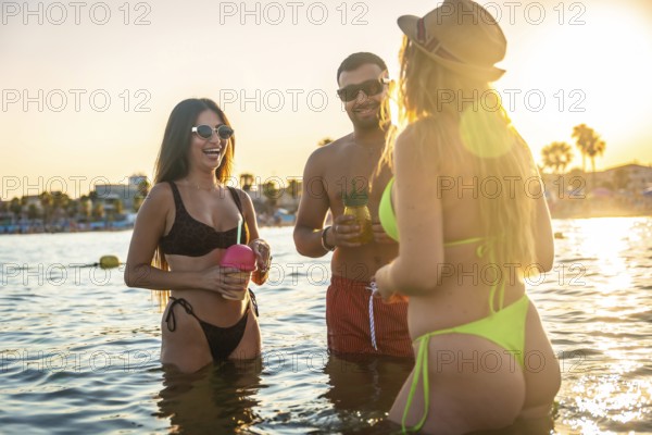Three friends enjoying refreshing cocktails while having fun in the ocean at sunset, embracing the