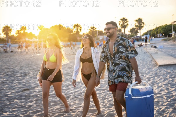 Three friends enjoying a leisurely stroll along the beach at sunset, carrying a cooler, ready for a