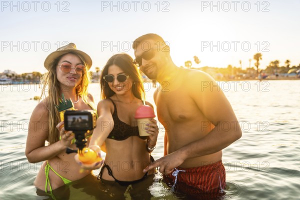 Three friends enjoying summer vacation, taking a selfie with action camera in the sea at sunset,