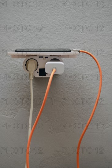A smartphone is plugged into a wall outlet, showcasing cables in orange and white. This setup