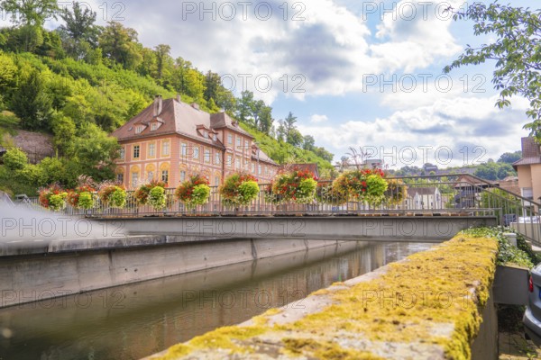 Historic building by the river, surrounded by blooming flowers and a green hill, small town pearl