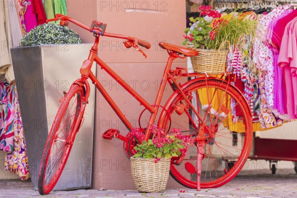 A decorated red bicycle stands in front of a clothing shop with colourful clothes, small town of