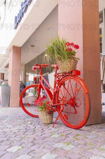 A red bicycle with flower baskets stands decoratively on a pavement in front of shops, small town