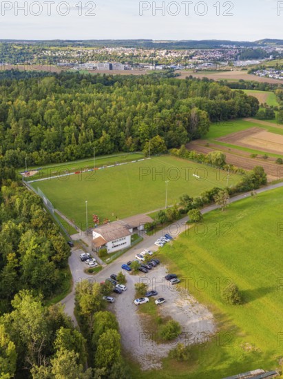 Aerial view of a sports field in the middle of wooded and rural surroundings with parked cars and a