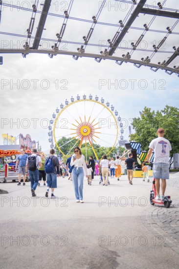 People in amusement park with e-scooter and big Ferris wheel in the background, Olympiapark,