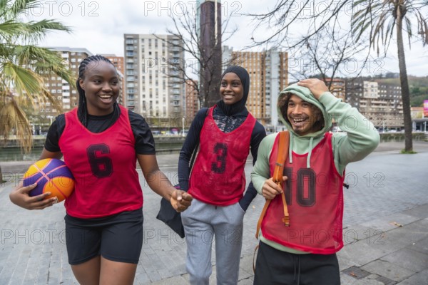 Three diverse friends, a young woman holding a basketball and two people wearing red bibs, walking