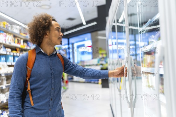 Young man with backpack opens supermarket freezer and selects products from a refrigerated aisle,