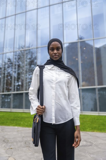 Young black muslim woman wearing a hijab and white shirt, holding a bag and standing confidently